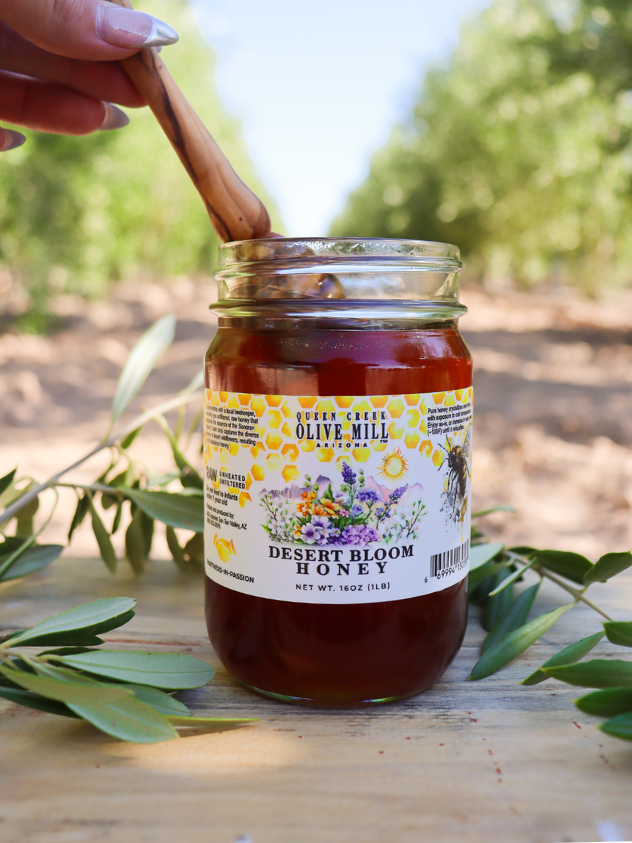 Jar of Queen Creek Olive Mill Desert Bloom Honey with a wooden spoon, surrounded by greenery
